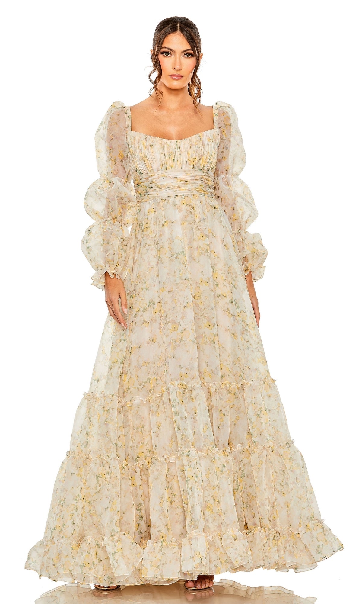 A woman models the Long Formal Dress 68288 by Mac Duggal—a flowing floral print gown with a ruched empire waist, puffed sleeves, and square neckline—standing against a white background.